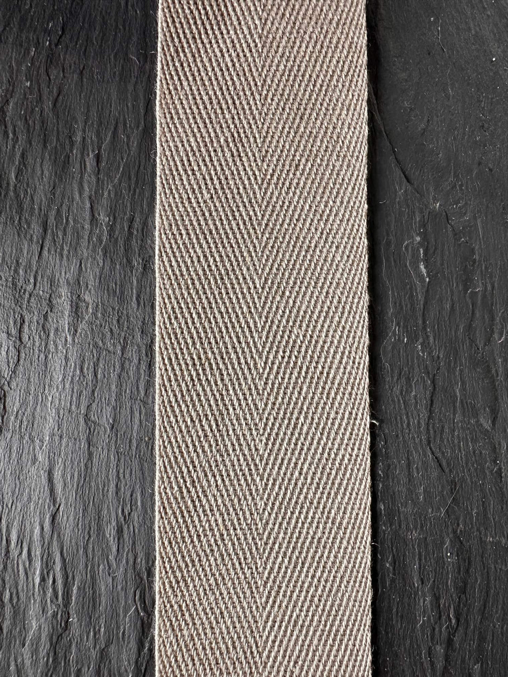 A close-up of a woven herringbone pattern hemp webbing tape  in a light beige or taupe color, positioned horizontally on a dark charcoal textured slate panel.